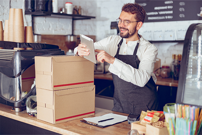 Café owner holding his tablet standing behind a counter with two cardboard boxes on top.