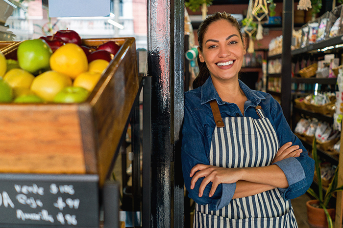 Woman in an apron smiling in the doorway of a grocery store with fresh produce on display in front of her.
