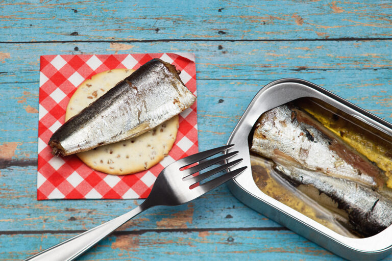 Opened tin of canned fish on a table beside a piece of fish on a cracker.