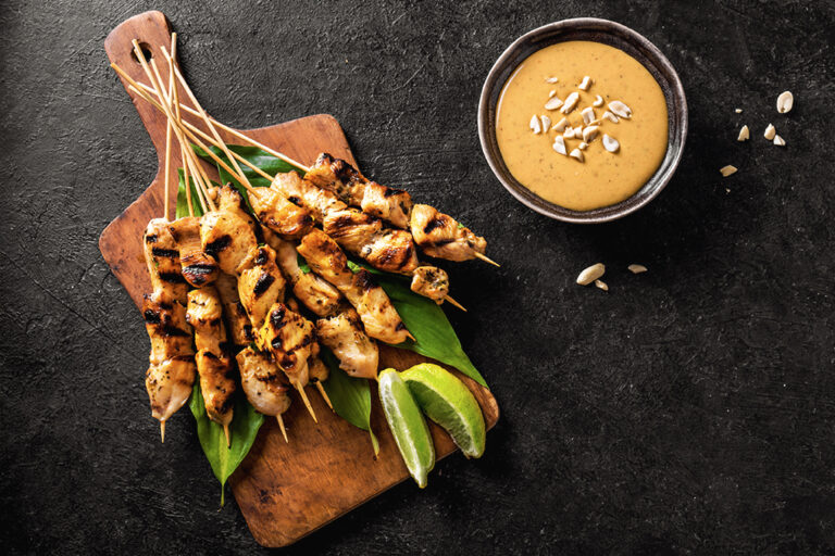 Brochettes de poulet servies sur une planche en bois avec des quartiers de lime, accompagnées d'un ramequin de sauce aux arachides.