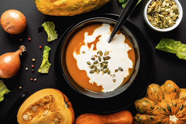 Un bol noir rempli de soupe crémeuse à la citrouille entouré d'une variété de légumes colorés.